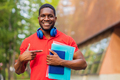 young afro american man with headphones on neck holding book and folder in hands in summer park - PhotoDune Item for Sale