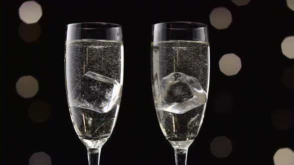 Two Filled Glasses of Champagne with an Ice Cube in Each. Bokeh Blinking Black Background. Close Up