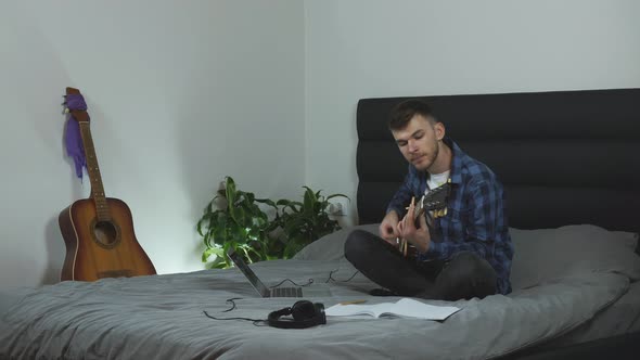 Man in pantone blue t-shirt plays on electric guitar sitting on bed at living room. Music concept