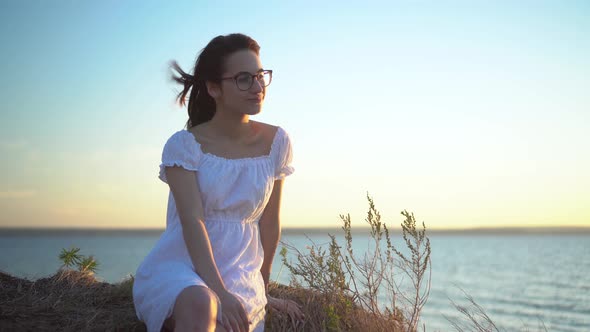 Attractive Young Woman Sits on a Hill Against the Background of the Sea. The Girl Is Resting in a