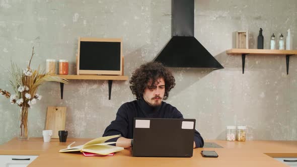 Serious Curlyhaired Young Man Working on Computer While Sitting in Kitchen