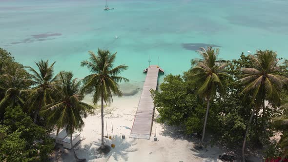 Drone flight over turquoise water to an island with green trees and a pier among palm trees