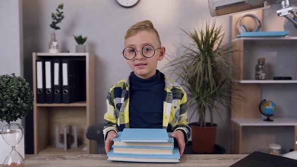 10-Aged Serious Boy in Glasses which Holding a Lot of Books on Table and Looking at Camera