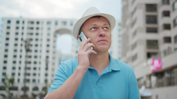 Young Man in White Hat Talking on a Mobile Phone While Standing on a City Street