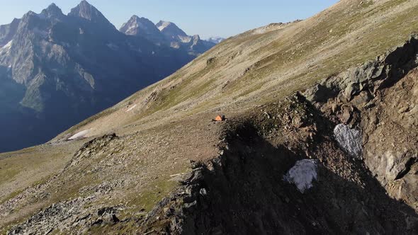 An Aerial View of an Orange Tent Stands on the Edge of a Cliff High in the Mountains on a Sunny Day