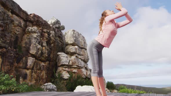Caucasian woman practicing yoga, stretching and touching floor, standing on deck on mountainside