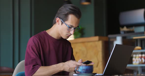 Young Man Drinking Coffee in a Cozy Cafe While Typing on Laptop and Chatting Using His Mobile Phone
