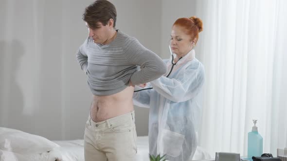 Ill Caucasian Man Standing in Hospital Ward with Concentrated Doctor Listening Back with Stethoscope