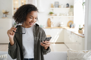 olding smartphone, shopping in online store at home. Modern teen lady with afro hairstyle chatting in social network, reading message with good news.
