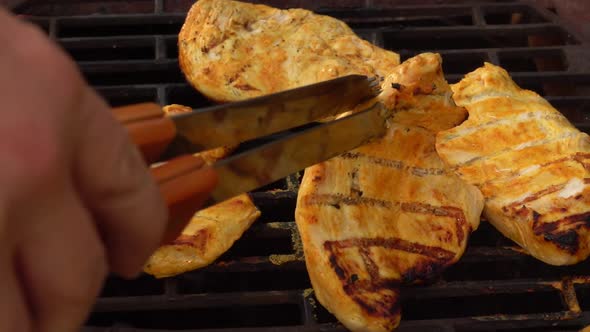 The Hand Is Checking Chicken Fillet on the Grill Grid with Kitchen Tongs