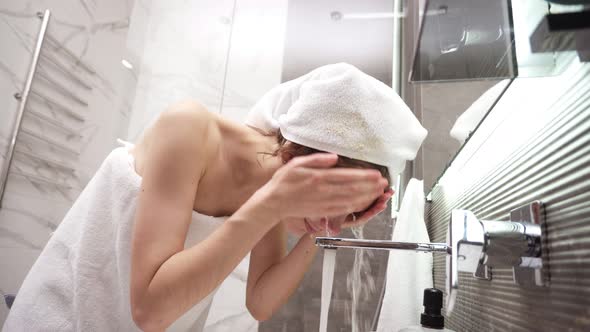 A Woman Standing in the Bathroom Near the Sink in White Towel on Head and Body Washes Her Face with