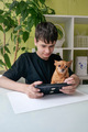 teenager sits at the table with his little dog and plays computer games on a tablet - PhotoDune Item for Sale