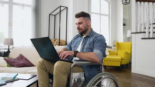 Modern Man with Beard Sitting in Wheelchair and Remotely Working at Home on Laptop