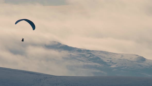 Paraglider shot against the light with snow covered hills and clouds in the background, Cumbria UK.