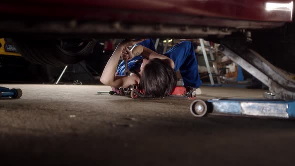 Woman Mechanic with Tattoos Lies Under Car on Car Repair Trolley and Repairs It with Tools