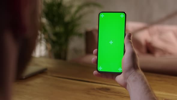 Handheld Camera Back View of Young Man at Home Sitting on a Wood Desk Using With Green Mockup Screen