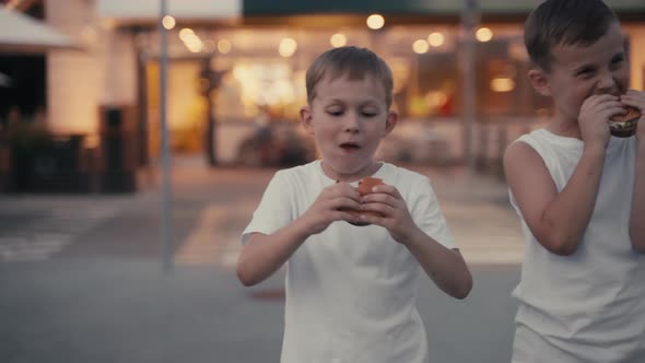 Two Hungry Young Boys Eating a Burgers on the Evening Street