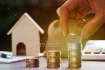Business and financial concept. A man hand putting coin on pile coins with burred small house, notebook, pen, money bag, calculator as background.