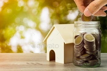 A businessman hand holding coin over glass jar and resident house model on wooden table.