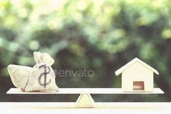  buying concept : Balance a money bag and small residential, house model on table against green nature background. Exchange of finances and houses.