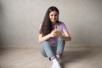 A young girl is sitting on the floor, smiling and holding a phone in her hands to take a photo