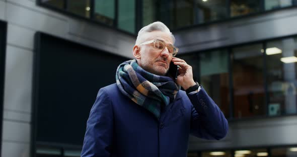 A Grayhaired Man Uses a Mobile Phone Standing in Business Center of the City