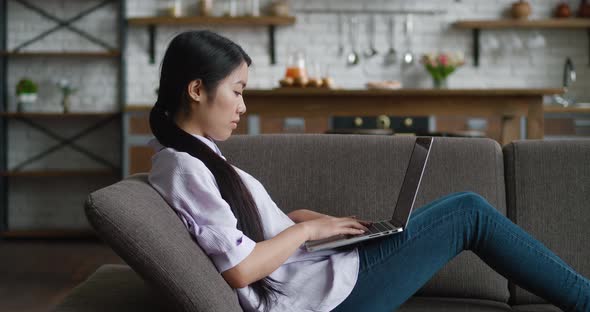 Asian Woman Resting on Couch Using Laptop Notebook Looking at Screen Typing Message at Living Room