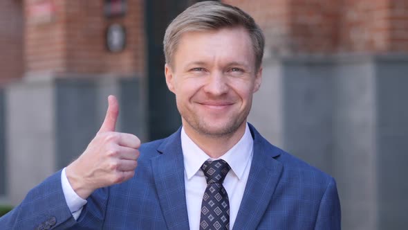 Thumbs Up By Businessman at Work Looking at Camera