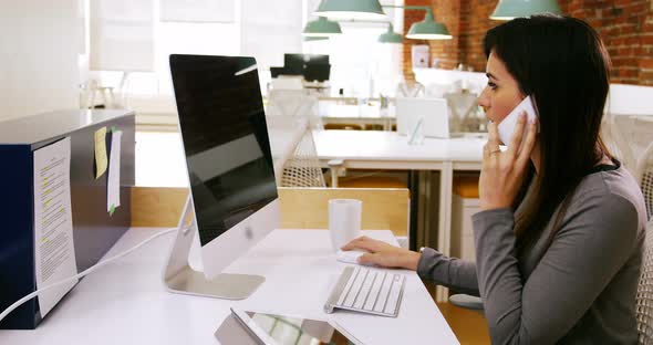 Female executive talking on mobile phone while working on computer at desk