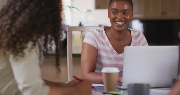 Two happy diverse female friends sitting in kitchen with coffee, working on laptop and laughing