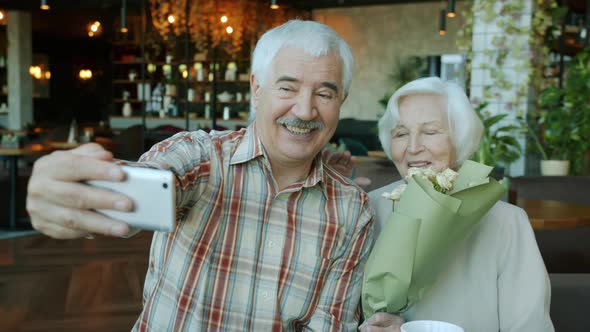 Senior Husband and Wife Using Smartphone To Take Selfie in Restaurant While Woman Holding Flowers