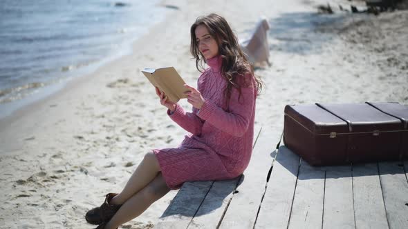 Wide Shot Absorbed Caucasian Woman Reading Book Sitting on Wooden Pier on Sandy Beach