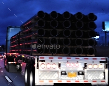 ing a flat bed trailer filled with large pipes, driving on the freeway at night.