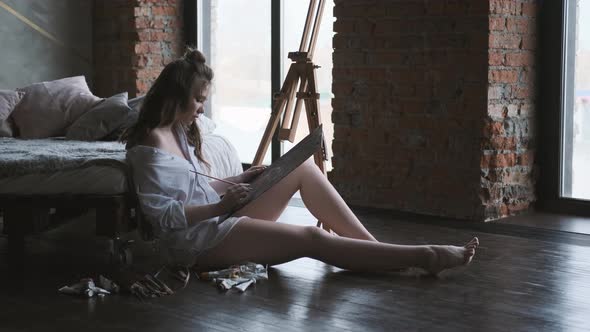 Painter Paints a Picture Sitting on the Floor in a Home Studio. Girl in a Man's Shirt