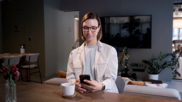 A beautiful girl with glasses sits at a table in the living room, looks into the phone.