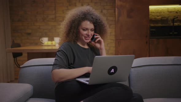 Young Woman Talking on Mobile Phone and Looking at Laptop Sitting on Couch at Home