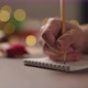Slow Motion Orbit Shot Man Writes Christmas Gifts Notepad Pencil Under Warm Light Evening - VideoHive Item for Sale