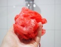 Hand holding red loofah with soap bubbles against white tiles in the bathroom. - PhotoDune Item for Sale