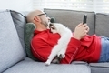 Man laying on the couch with mobile phone and kitty playing on his chest. - PhotoDune Item for Sale