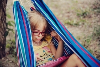 Cute little girl in eyeglasses laying in a hammock and using tablet.