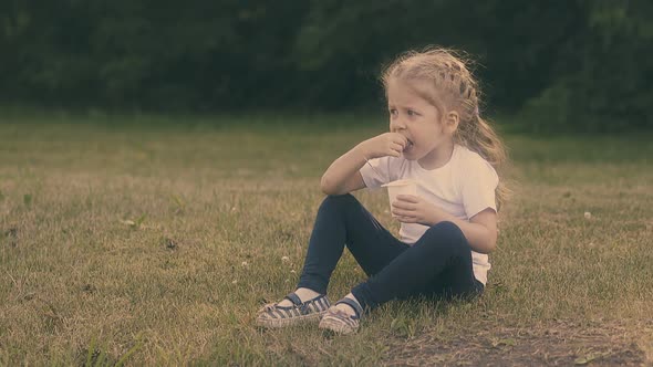 Little Pretty Lady with Fair Hair Eats Yogurt on Park Grass