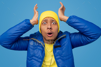 eeping hands near head and looking at camera in disbelief against blue background