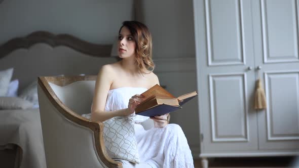 Woman in white dress sitting in armchair in vintage interior and reading a book