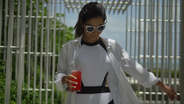 Happy Young Lady in White Shirt Walking on Sun Terrace of Tropical Hotel Holding Glass of Red