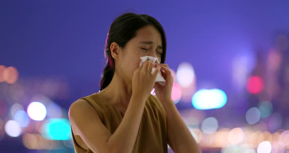 Woman sneezing with tissue in city at night