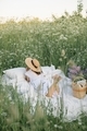 Woman in dress and straw hat relaxing on white bed linen in the field with tall grass and flowers. - PhotoDune Item for Sale