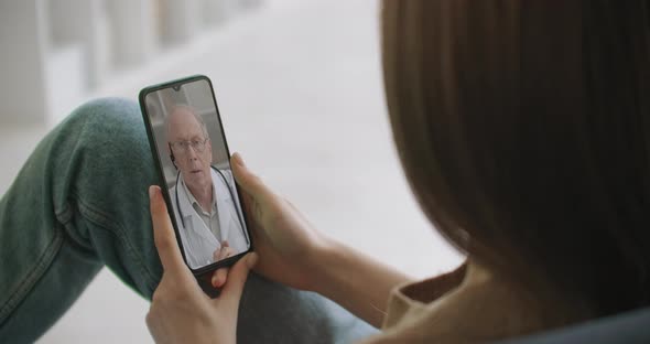 Female Medical Assistant Wears White Coat, Headset Video Calling Distant Patient on Smartphone