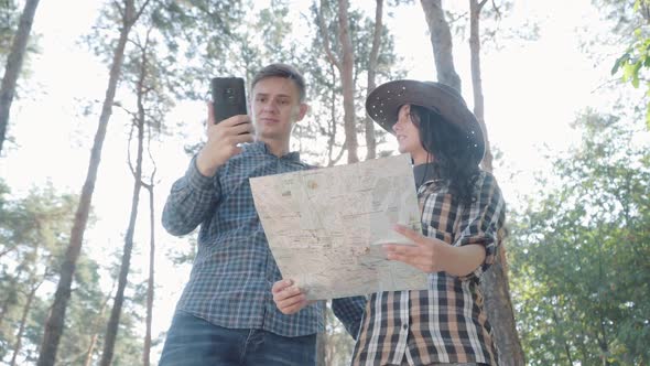 Young Couple Checking Paper and Online Map in Summer Forest. Busy Caucasian Man Answering Phone Call