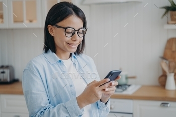 pps on phone, shopping in online store, standing in kitchen at home. Smiling young woman uses smartphone, checking news, chatting in social networks.