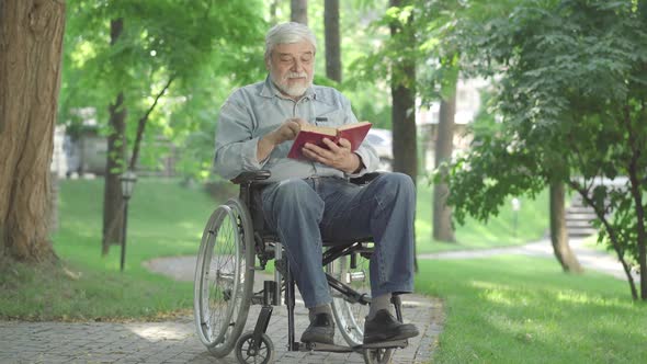 Disabled Senior Man Sitting in Wheelchair and Reading Book. Wide Shot Portrait of Positive Caucasian